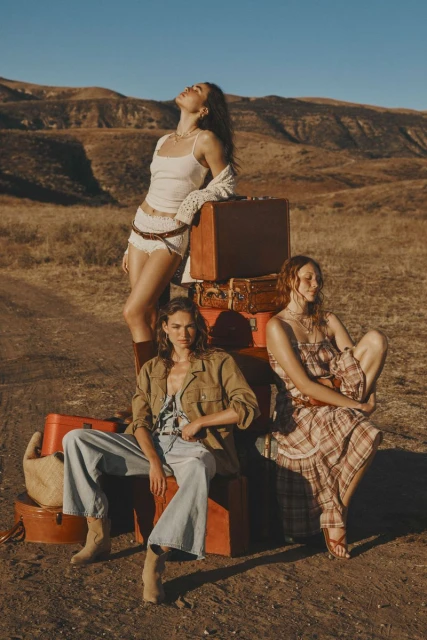 Three women in casual fashion, posing near vintage luggage in a dry, hilly landscape with a clear blue sky in an outdoor fashion editorial.