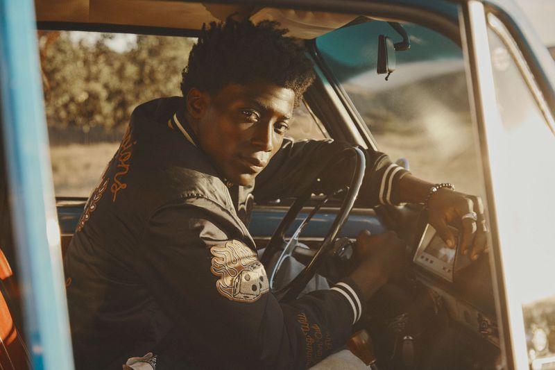 An adult Black male sits in a vintage blue car with a varsity jacket. His hand is on the steering wheel and radio.