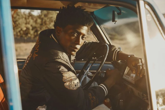 An adult Black male sits in a vintage blue car with a varsity jacket. His hand is on the steering wheel and radio.