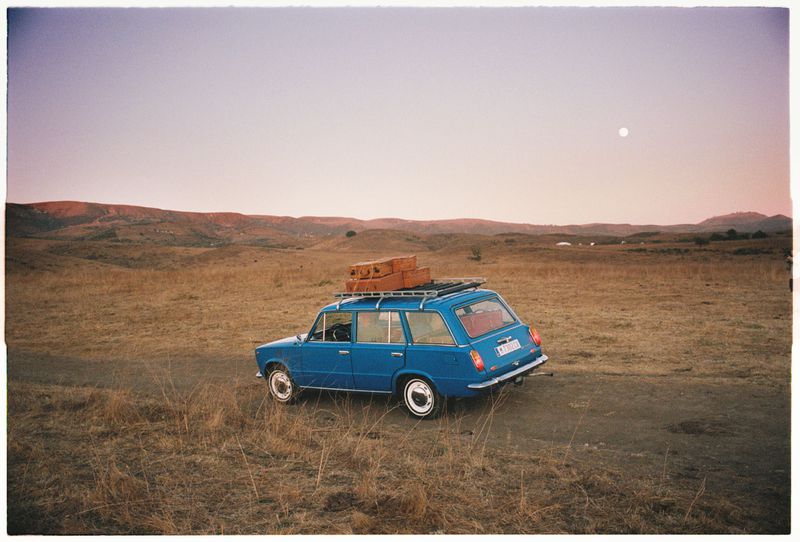 Vintage blue station wagon parked in a dry field under a pale sky with luggage strapped to the roof rack. Rural scenery in background.