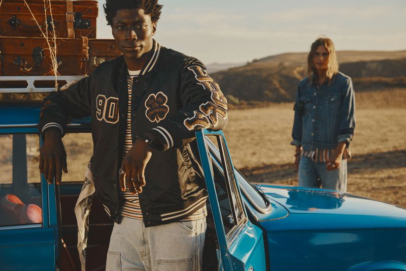 Two adult humans outdoors with a vintage blue car and stacked luggage on top. Man in bomber jacket leans on the car, another stands nearby.