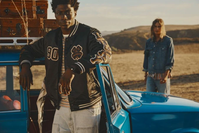 Two adult humans outdoors with a vintage blue car and stacked luggage on top. Man in bomber jacket leans on the car, another stands nearby.