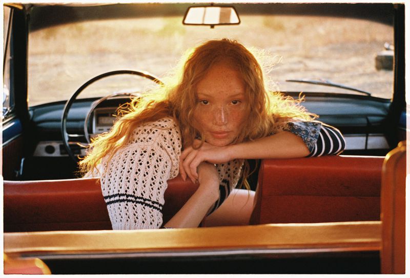 A young woman with red hair and freckles leans against the red backseat of a vintage car with a dark blue exterior, bathed in warm light.
