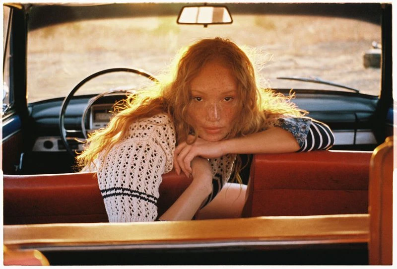 A young woman with red hair and freckles leans against the red backseat of a vintage car with a dark blue exterior, bathed in warm light.