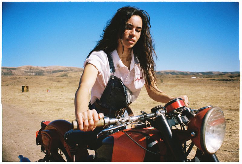 A brunette woman on a red motorcycle in a rural landscape under a clear blue sky, wearing a white shirt and denim overalls.