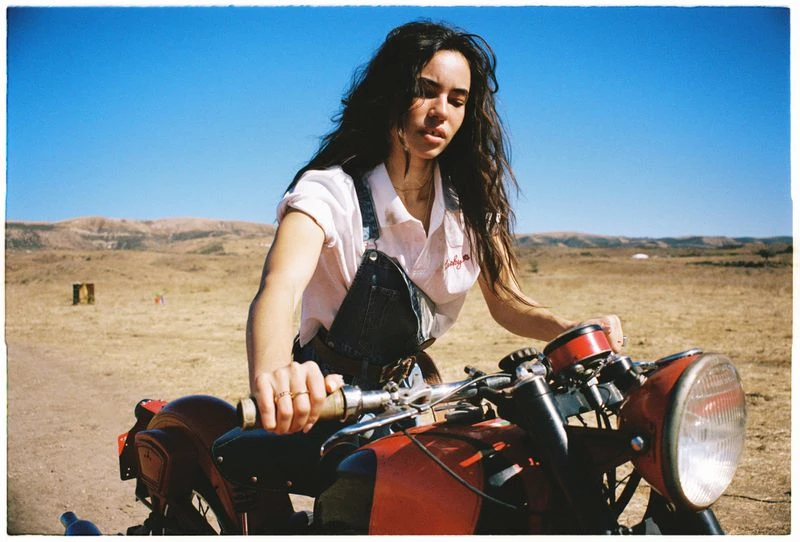 A brunette woman on a red motorcycle in a rural landscape under a clear blue sky, wearing a white shirt and denim overalls.