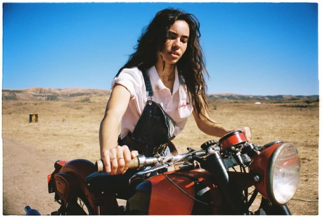A brunette woman on a red motorcycle in a rural landscape under a clear blue sky, wearing a white shirt and denim overalls.