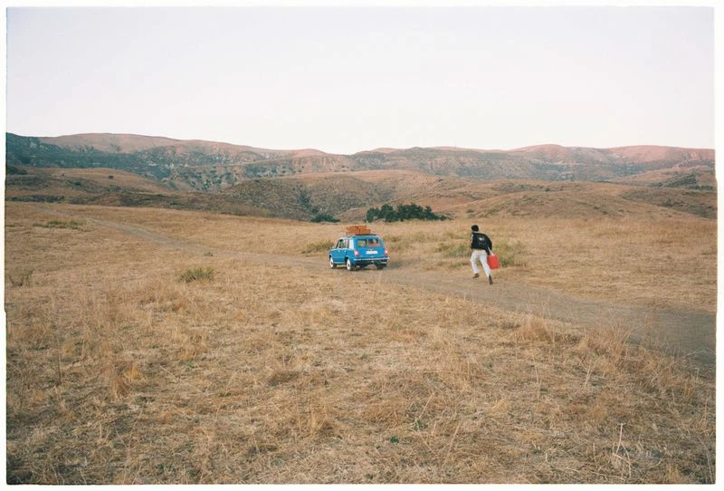 Blue car with luggage on top travels a dirt road near a person carrying a red container, set against a hilly, grassy landscape.