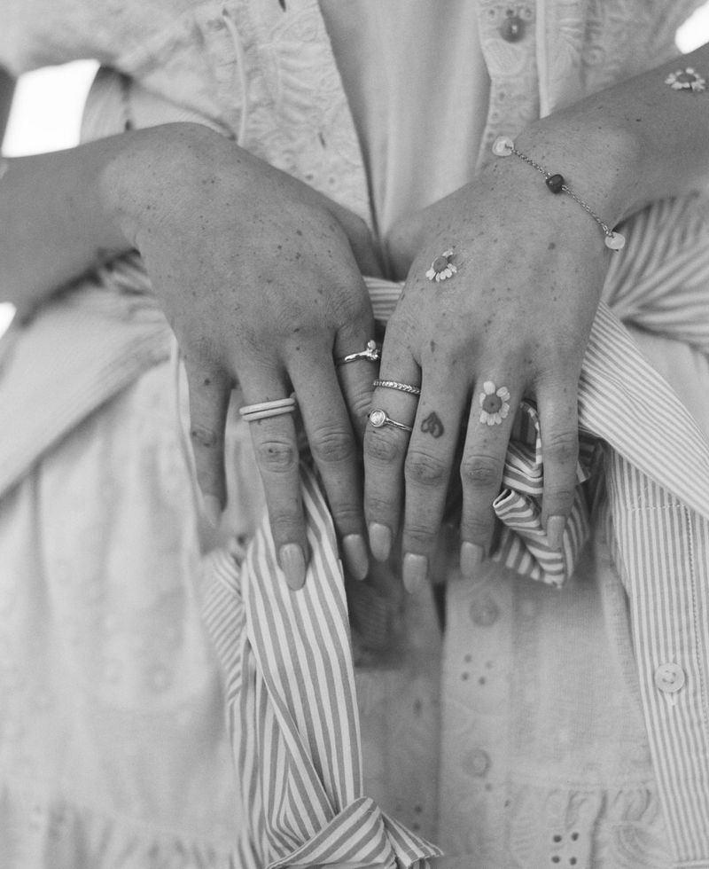 Close-up of a person's hands with rings, bracelets, and small flower tattoos, holding a striped garment, set against a neutral background.