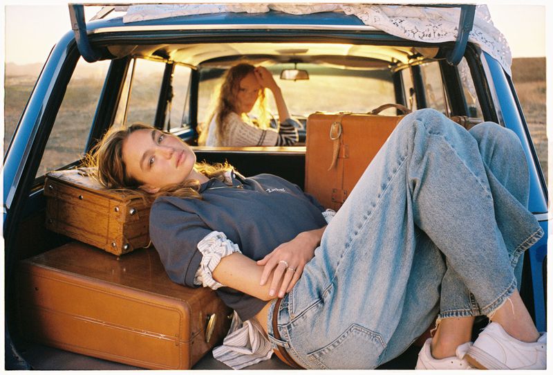 Two women in a vintage car with luggage, enjoying a road trip in golden sunlight, casual clothing, and natural hair.