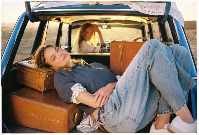 Two women in a vintage car with luggage, enjoying a road trip in golden sunlight, casual clothing, and natural hair.