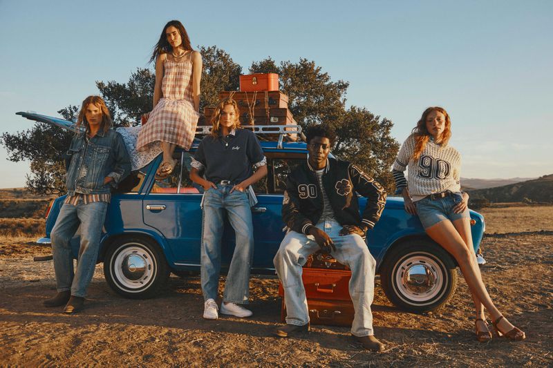 Five fashion models pose beside a blue car in a sunlit field, wearing casual clothing and surrounded by trees and vintage suitcases.