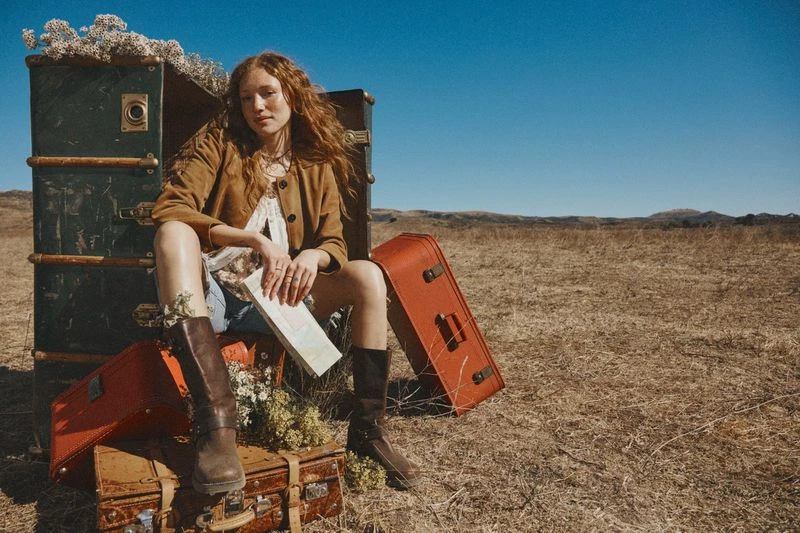 A woman with auburn hair sits on vintage luggage in a field under a blue sky wearing a jacket and boots in an outdoor shot.