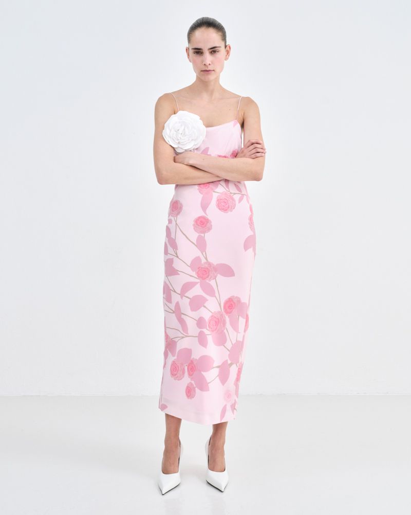 Elegant adult woman in a pink floral dress and white heels posing with her arms crossed in a clean, bright photo studio setting.