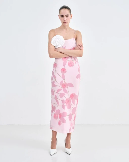 Elegant adult woman in a pink floral dress and white heels posing with her arms crossed in a clean, bright photo studio setting.