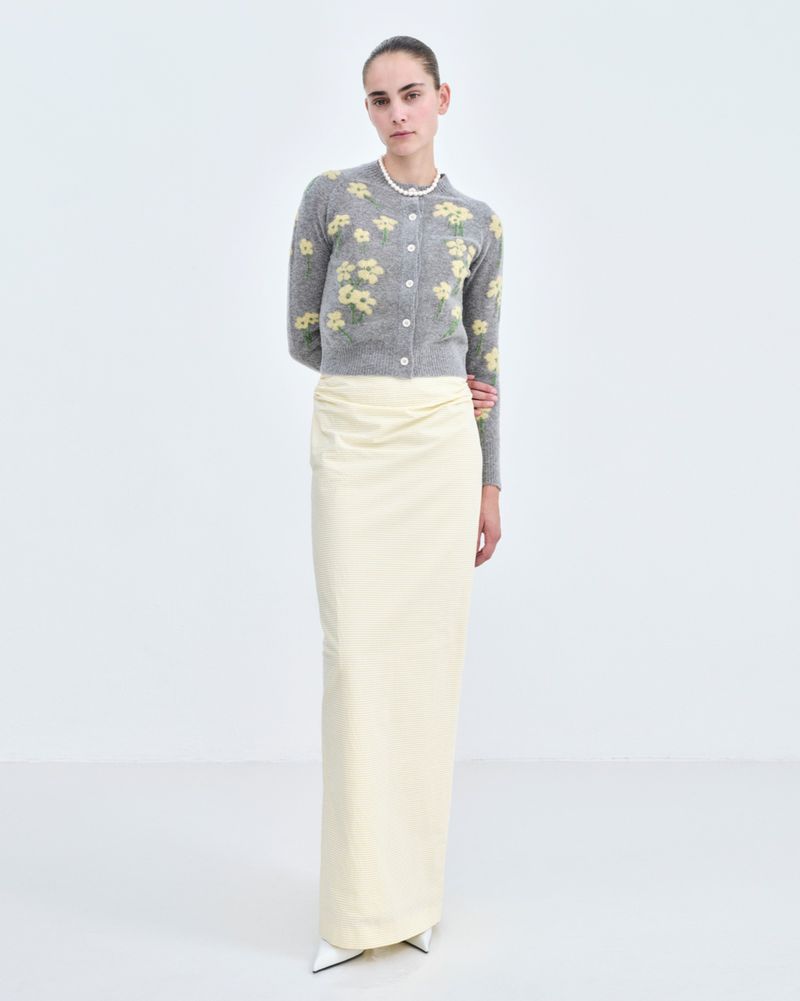 Fashion model poses in a light gray floral cardigan and long white skirt in a white photo studio setting. Full-length shot, eye-level angle.