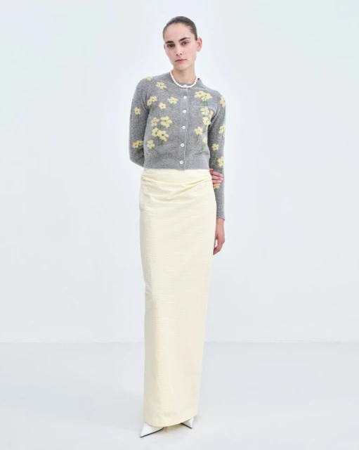 Fashion model poses in a light gray floral cardigan and long white skirt in a white photo studio setting. Full-length shot, eye-level angle.