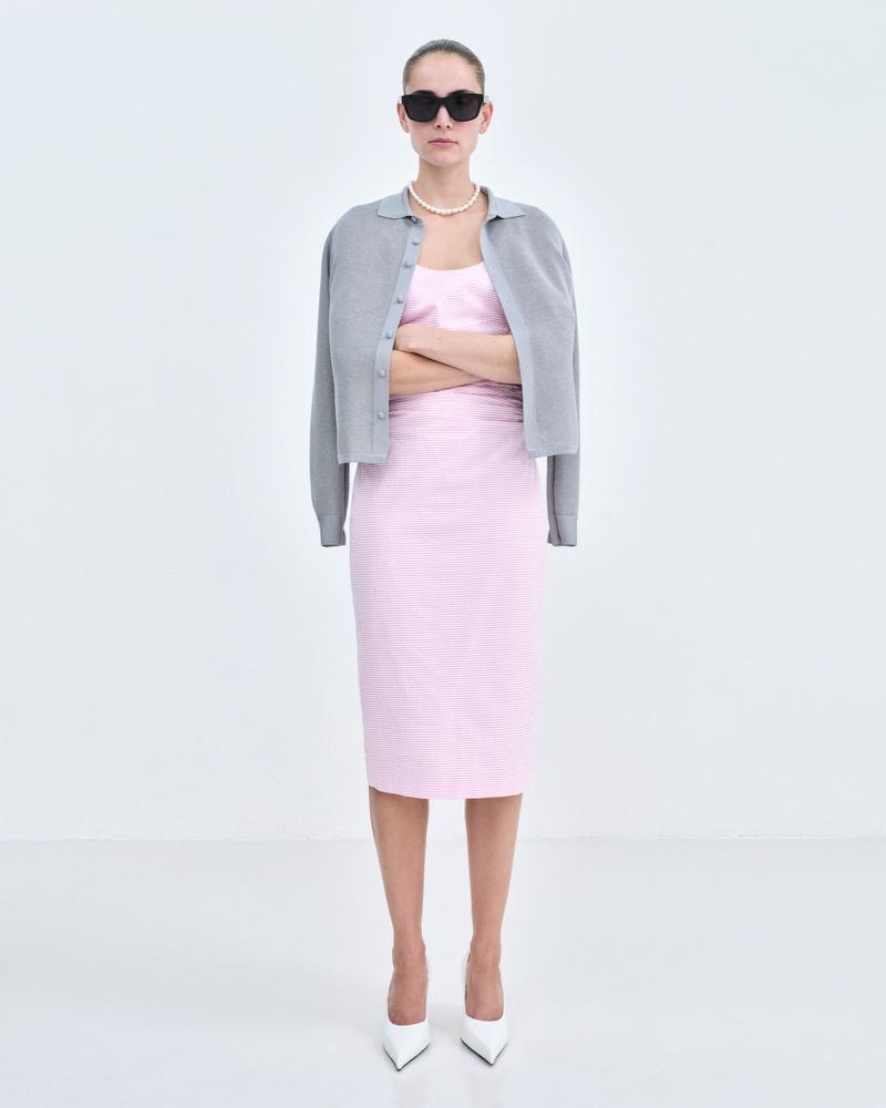 Full shot of an adult woman in a light pink dress and gray cardigan with white heels and sunglasses, standing against a white background.