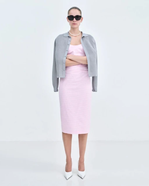 Full shot of an adult woman in a light pink dress and gray cardigan with white heels and sunglasses, standing against a white background.