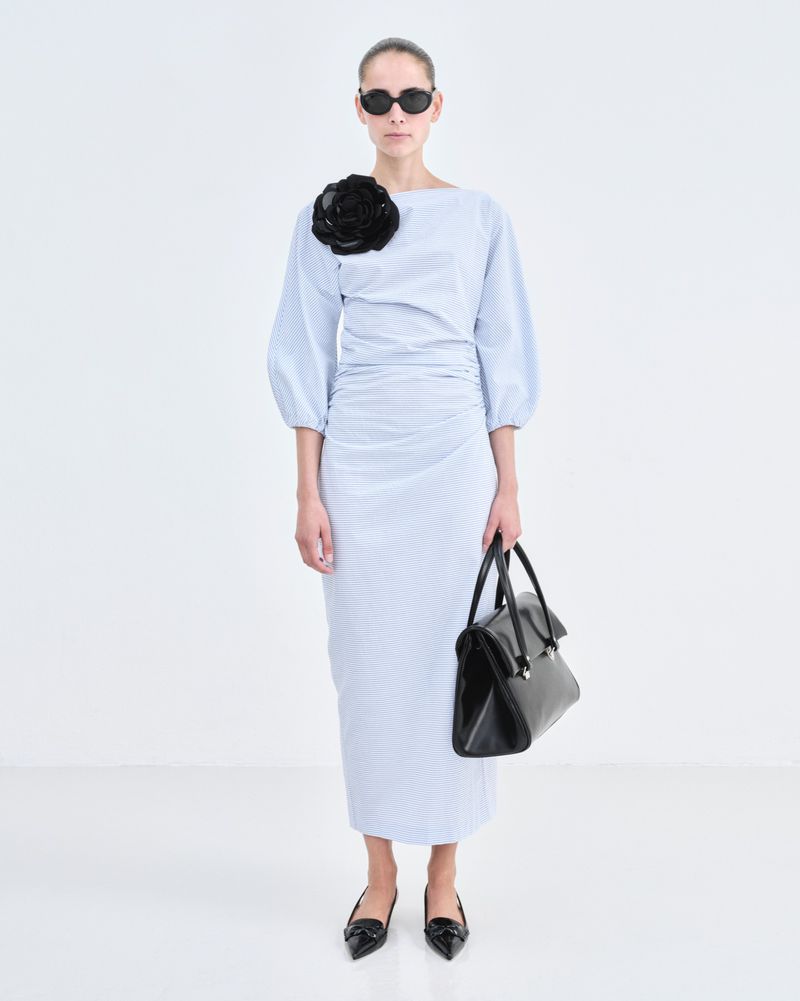A woman in a light blue dress with a black flower, sunglasses, and a black bag stands in a white photo studio, showcasing a modern fashion look.