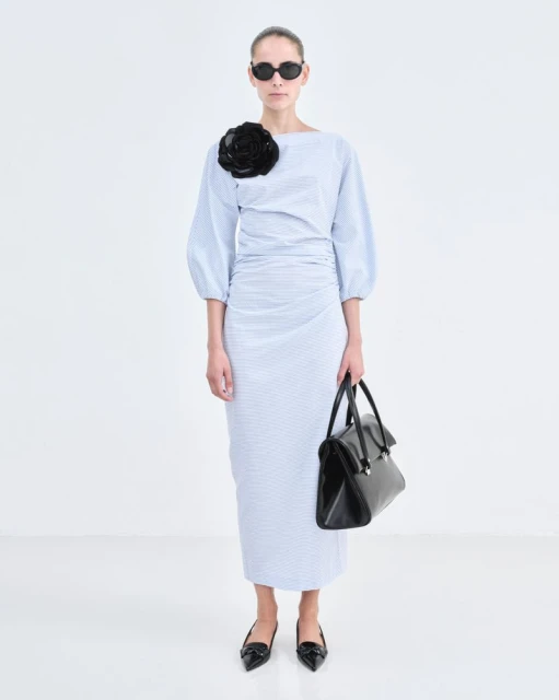 A woman in a light blue dress with a black flower, sunglasses, and a black bag stands in a white photo studio, showcasing a modern fashion look.
