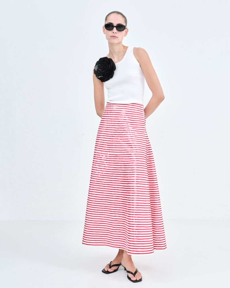 Woman in a white top and a red and white striped skirt poses in a white studio. She is wearing black sunglasses and flip flops.