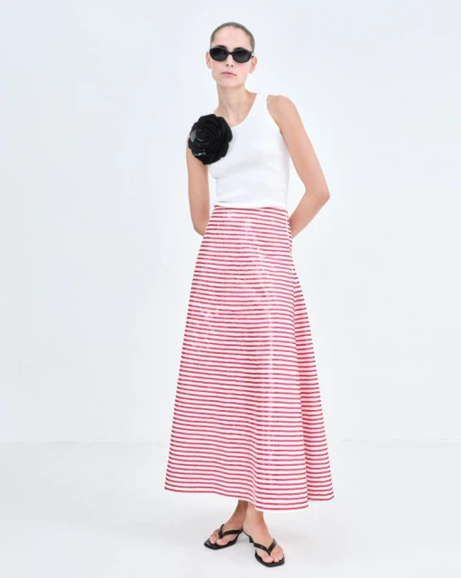 Woman in a white top and a red and white striped skirt poses in a white studio. She is wearing black sunglasses and flip flops.