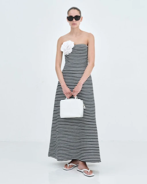 An adult female model poses in a striped strapless dress with a white flower detail, holding a white purse in a bright studio.