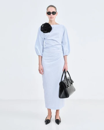 Model Romy Schonberger wearing a blue dress with a black flower applique and holding a black handbag against white seamless