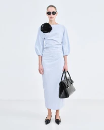 Model Romy Schonberger wearing a blue dress with a black flower applique and holding a black handbag against white seamless