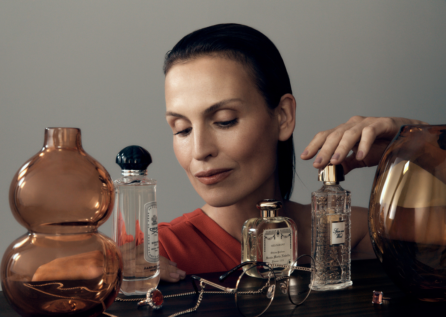 Elegant adult woman posing with an array of vintage-style perfume bottles and jewelry in a softly lit, neutral studio setting.
