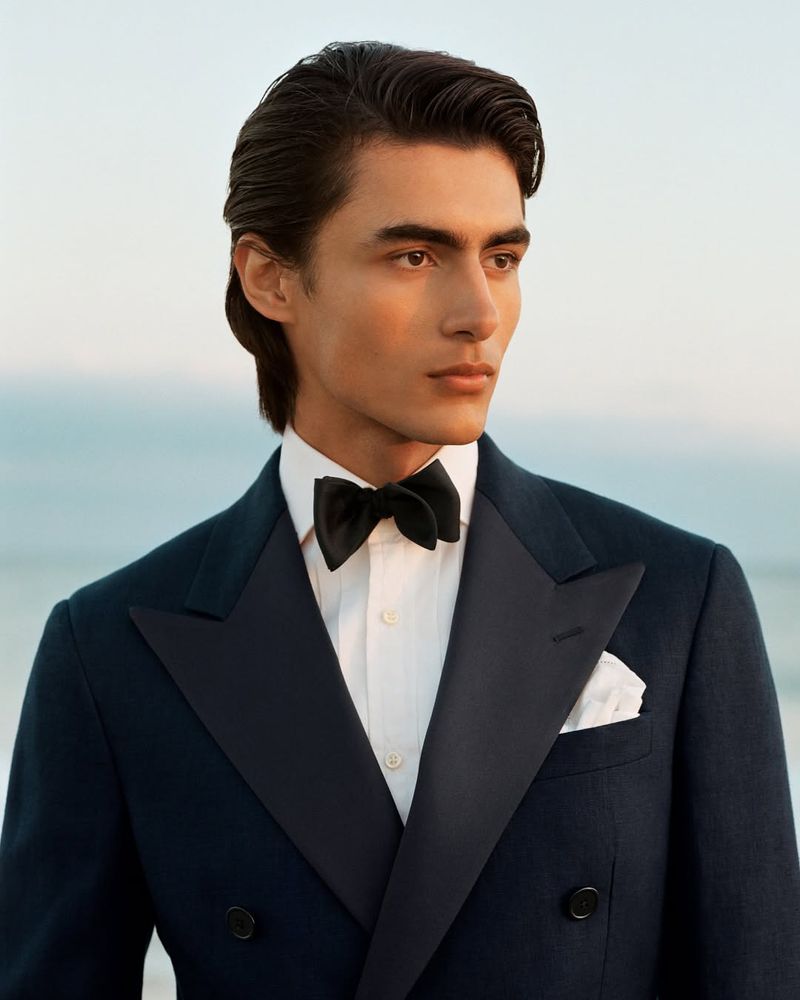 Portrait of a man in a suit and bow tie, gazing into the distance against a muted seascape backdrop with soft, diffused lighting.