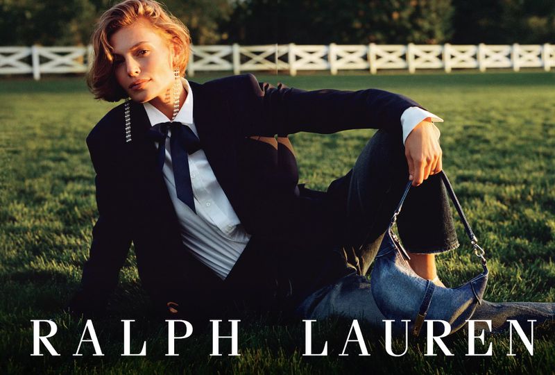Elegant woman in Ralph Lauren blazer, bow tie, and jeans lies on green grass with denim purse, posing against a backdrop of trees and white fence.