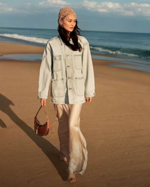 Adult woman walking on beach wearing a crocheted beanie, light blue denim jacket, and tan satin dress, carrying a brown purse.