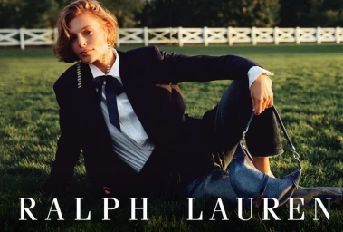 Model seated on grass wearing a dark blazer, white shirt with bow tie, and denim bag for Ralph Lauren.