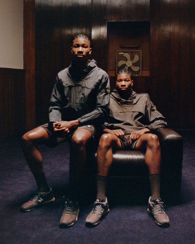 Two young Black male models are seated indoors in a studio, wearing grey jackets and shorts, and sneakers with socks. 