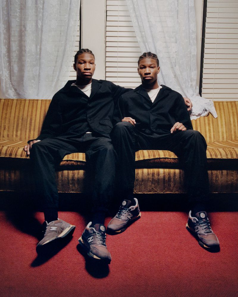 Two young adult men in dark outfits sit on a gold couch with shoes in front of them, inside a home or photo studio setting.