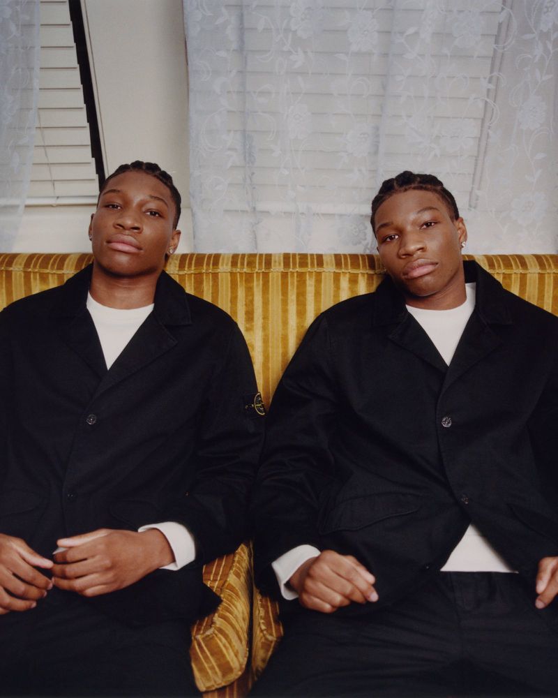 Portrait of two adult males with braids, wearing black jackets, sitting on a yellow couch in an indoor setting with a white curtain.