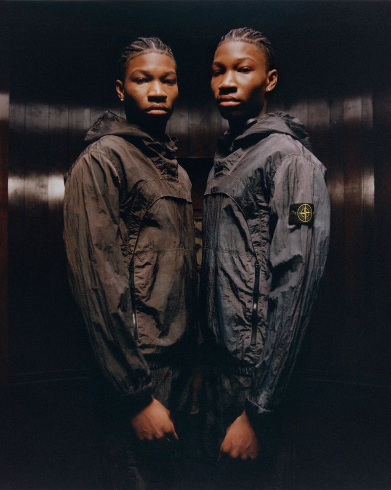 Two young Black men pose indoors wearing matching hooded jackets; fashion shot with dramatic lighting and neutral tones highlighting details.