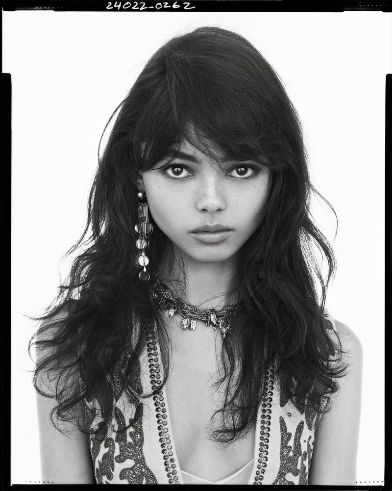 Black and white portrait of a woman with dark wavy hair wearing long statement earrings against a white background.