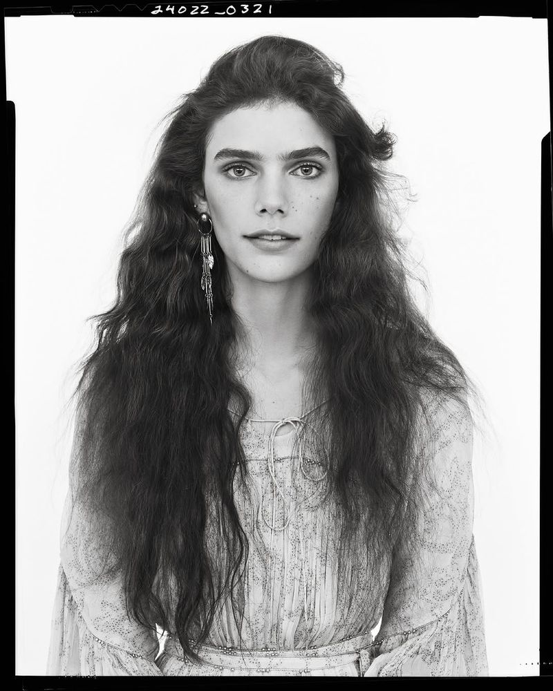 Portrait of a woman with long, wavy brown hair wearing a light-colored, patterned top and long, dangling earrings, set against a white backdrop.