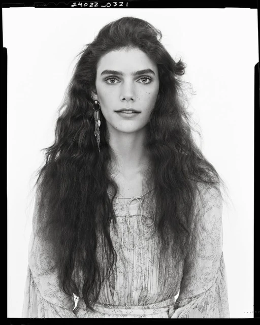 Portrait of a woman with long, wavy brown hair wearing a light-colored, patterned top and long, dangling earrings, set against a white backdrop.
