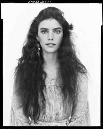 Portrait of a woman with long, wavy brown hair wearing a light-colored, patterned top and long, dangling earrings, set against a white backdrop.