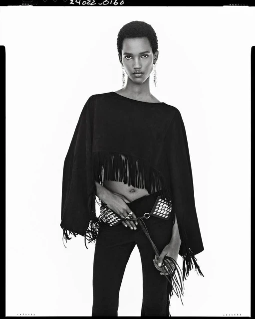 A black and white fashion portrait of a model wearing a fringed black poncho top, studded belt, and dark pants against a white backdrop.