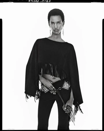 A black and white fashion portrait of a model wearing a fringed black poncho top, studded belt, and dark pants against a white backdrop.
