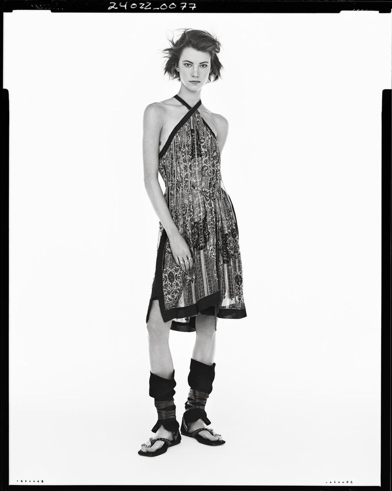Black-and-white portrait of a young adult female model in a halter dress, sandals, and leg warmers against a white background.