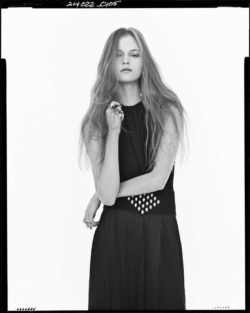 Portrait of a young woman with long wavy hair posing in a sleeveless black dress in a brightly lit studio. Fashion editorial.