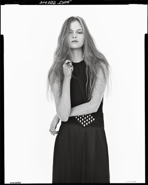 Portrait of a young woman with long wavy hair posing in a sleeveless black dress in a brightly lit studio. Fashion editorial.