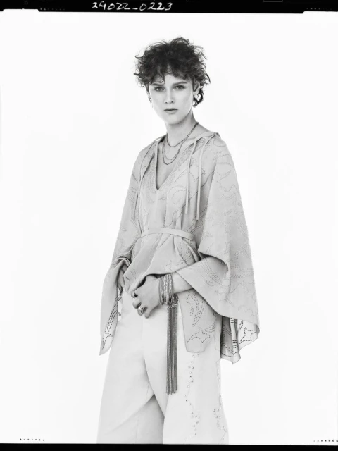 A woman with curly short brown hair wears a light-colored hooded poncho and white pants in a neutral studio environment. She's posing.