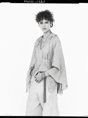 A woman with curly short brown hair wears a light-colored hooded poncho and white pants in a neutral studio environment. She's posing.